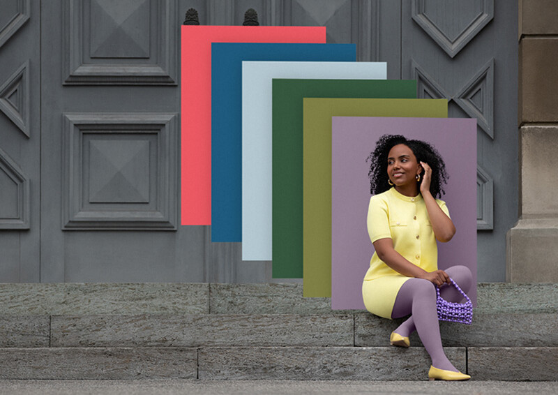 Juzo Trend Colours Batik Una donna è seduta sui gradini di pietra davanti a un cancello. Indossa un completo due pezzi giallo chiaro abbinato a un bendaggio compressivo viola. Dietro di lei si vedono campioni di colore nei colori di tendenza