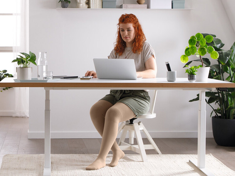 Eine Frau sitzt an einem Schreibtisch an ihrem Laptop, sie trägt Juzo Expert Flachstrickkompressionsstrümpfe