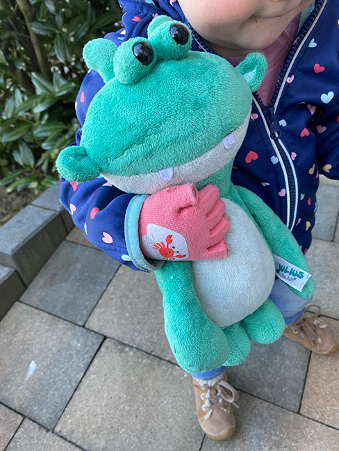 Clara mit Julius ein kleines Mädchen trägt an der Hand einen rosanen Kompressionshandschuh und hält ihr Julius Kuscheltier fest