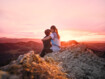 Eine Mutter sitzt mit ihrem Kind im Arm auf einem Berggipfel und betrachtet den Sonnenuntergang