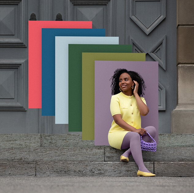A woman sits on stone steps in front of a gate. She wears a light yellow two-piece outfit with a purple compression garment. Behind her, you can see colour fields in the trend colours.