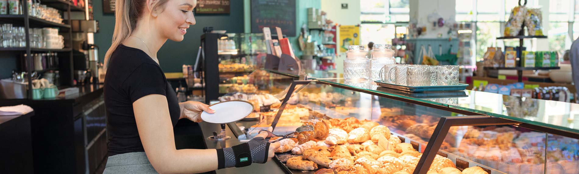 Lifestyle-billede bagerforretning - JuzoFlex Manu Xtra Ekspedient i bagerforretning bærer håndledsbandagen JuzoFlex Manu Xtra og tager en croissant op med en serveringstang