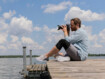 Mann sitzt auf einem Steg am See und fotografiert mir seiner Kamera. Am Knie trägt er eine Bandage