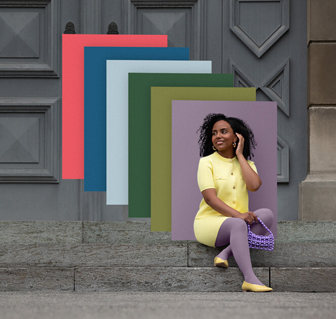 Una mujer está sentada en unos escalones de piedra frente a una puerta. Lleva un conjunto de dos piezas de color amarillo claro y una compresión de color lila. Detrás de ella se ven campos de color en los colores de moda.