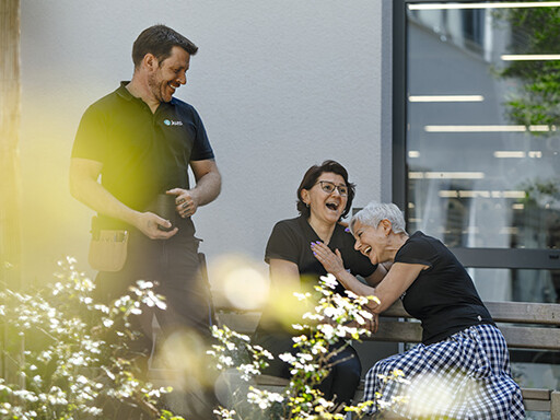 Unsere Pausen verbringen wir gerne gemeinsam im Freien Zwei Frauen mit einem schwarzen Juzo T-Shirt sitzen auf einer Bank und lachen herzhaft, sie unterhalten sich mit einem Mann, der ebenfalls ein schwarzes Juzo T-Shirt trägt und eine schwarze Tasse in der Hand hält
