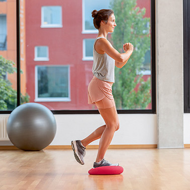 Uebung Einbeinige Squats Schritt 1 Frau steht mit einem Bein auf einem weichen Kissen und hebt das andere Bein leicht an