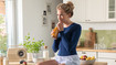 Una mujer con un vendaje de tórax azul oscuro está sentada en una encimera de cocina y bebe un zumo de naranja.