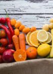 Auf einem holzigen Hintergrund erkennt man buntes Obst und Gemüse aufgereiht nebeneinander. Es bildet sich so ein Farbverlauf von rot, orange, gelb bis zu grün und rot