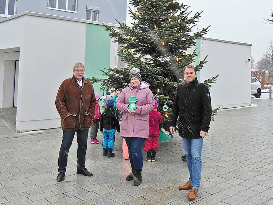 Wir unterstützen den Julius-Kindergarten Zwei Männer und eine Frau mit einem Julius-Plüschtier stehen draußen vor einem großen Tannenbaum, um den sich Kinder vom Julius Kindergarten versammelt haben