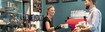 Verkoopster in bakkerij draagt JuzoFlex Manu Xtra polsbandage en reikt klanten over de toonbank een kopje aan