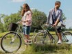 Eine Frau schiebt ihr Fahrrad neben einem Mann, der auf einem Fahrrad sitzt, im Hintergrund ist ein See zu sehen und Bäume