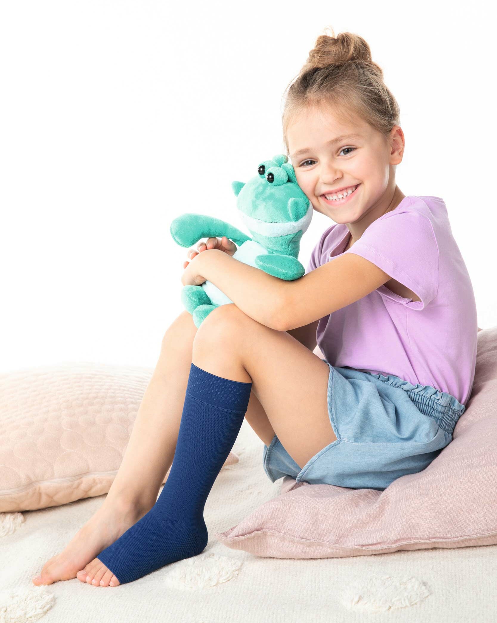 Ein blondes Mädchen sitzt auf einem Kissen, am linken Bein eine blaue Kompressionsversorgung und hält ein Julius Kuscheltier im Arm